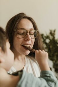 A woman with eyeglasses enjoys chocolate fed by a child at home.
